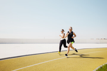 Two women joyfully jogging under a bright sun on a vibrant track amidst a clear blue sky in a...
