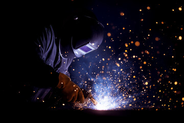 Welder engages in welding process, sparks erupting from welding torch in dimly lit workshop. Creative metalworking setting, highlighting skill and precision