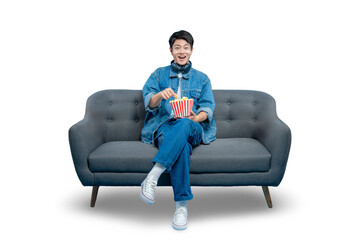 A focused young man eats popcorn from a bucket while sitting on a sofa, engrossed in a movie or...