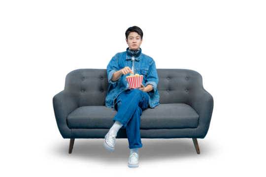 A focused young man eats popcorn from a bucket while sitting on a sofa, engrossed in a movie or show.PNG