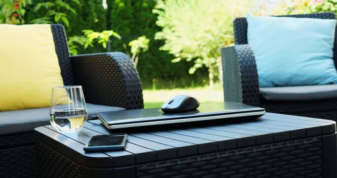 Modern devices laptop and smartphone left on small wooden table on terrace at armchairs against garden. Home summer terrace allows to spend free time in nature