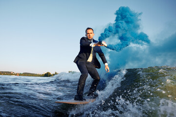 Young man in classic suit with blue smoke bomb in his hand rides wakeboard on a lake near city....