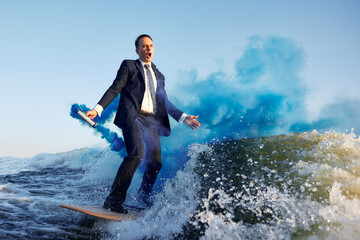 Young man surfer in classic suit with blue smoke bomb in his hand rides on the wave. Happy clerk...