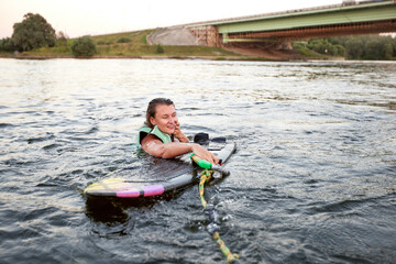 Young beautiful sporty girl in a green life jacket swims in the water with wakeboard and halyard in...