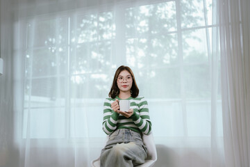 A young woman in a striped shirt and glasses relaxes indoors, enjoying music or a podcast through...