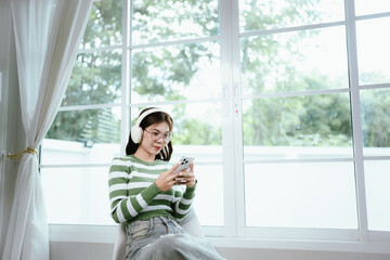 A young woman in a striped shirt and glasses relaxes indoors, enjoying music or a podcast through...