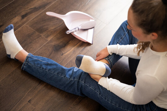 Woman looking at her dirty socks on feet after household cleaning, sitting in living room on wooden floor next to small plastic brush and dustpan, tired female resting after chores sweeping, top view
