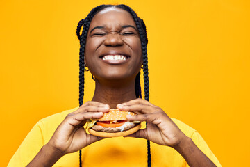 Happy woman holding a delicious burger in front of a vibrant yellow background, showcasing her joy...