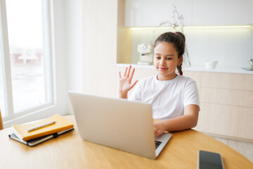 Young girl engaging in an online video call at home during the afternoon, smiling and waving...