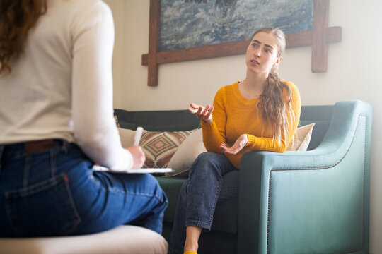 Young woman talking to psychologist during therapy session, sitting on sofa, female sharing problems while psychologist listening to her, mental health support, counseling in cozy modern interior