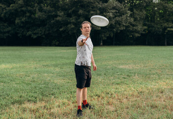 Focused male athlete in patterned white shirt, black shorts, smartwatch extends arm releasing white flying disc toward viewer over spacious grassy field, showcasing active sporty outdoor summer lifest