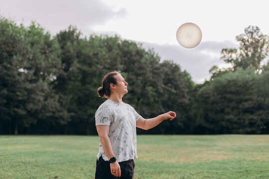 Athletic young adult with tied hair focused on frisbee disc floating midair green lawn background trees overcast sky modern casual wear expressive stance outdoor leisure activity