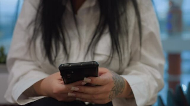 Close up of woman operating phone with polished nails and visible tattoo on right hand, wearing white shirt, background features soft blur with hints of vehicles in motion through large glass windows
