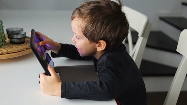 Happy boy presses on screen of tablet with finger playing interesting game. Curious little kid comments engaging funny app for children in room - Powered by Adobe