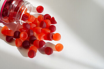 A transparent bottle lies on its side as colorful red and orange gummy vitamins spill onto a clean...