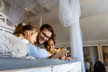 Excited single father plays with diverse, multi-ethnic daughters. Using digital tablet enjoy weekend