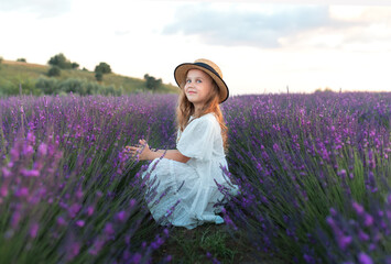 Portrait cute child girl sits in the middle of lavender bushes. Provence. Beautiful young girl in straw boater hat and a white dress collects lavender on lavender field.	
