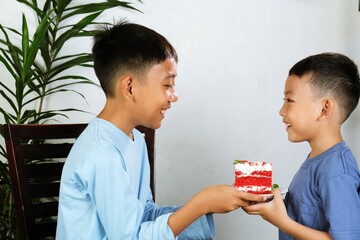 Two cheerful Asian boys enjoying a moment together while sharing a slice of red velvet cake at home with smiles and joy.