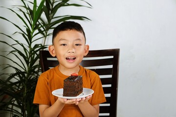 Smiling Asian boy holding a plate of chocolate cake with strawberry topping. Perfect for food, celebration, and childhood joy concepts.