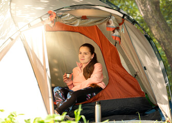 Young female traveler holding a stainless mug in a tent, enjoying the calm morning atmosphere in the forest during a solo camping trip.