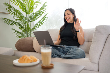 Young woman waving during a video call on laptop while sitting on a sofa at home.