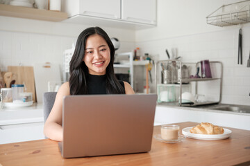 Confident young woman smiling at the camera while working on a laptop from her kitchen table with coffee and pastry on the side.