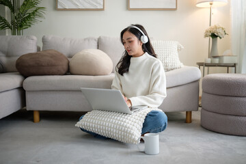 Young Asian woman wearing headphones, working on a laptop while sitting cross-legged on the floor...