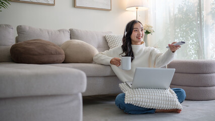 Casual lifestyle moment of a smiling woman multitasking with laptop, coffee, and TV remote.
