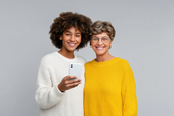 Young girl looking at camera while holding smartphone beside smiling senior woman. Bright neutral background highlights their joyful expressions. Concept of family bonding, technology, connections
