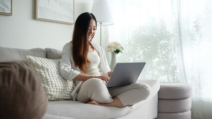 Young woman sitting comfortably on a sofa working on her laptop in a bright living room.