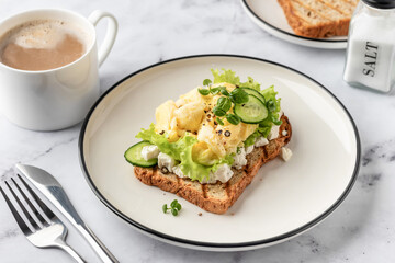 Scrambled eggs on grilled toast with salad leaves, cheese and micro greens on plate on white marble...