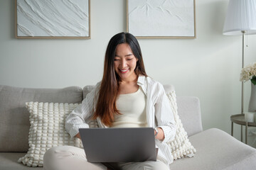 Young woman sitting comfortably on a sofa working on her laptop in a bright living room.