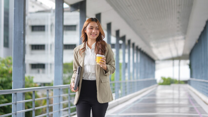 Fototapeta premium Confident young businesswoman walking across an office walkway with a coffee cup and a laptop in hand.