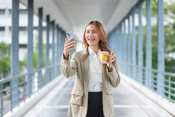 Smiling Asian businesswoman holding coffee and using her phone outdoors in an urban environment.