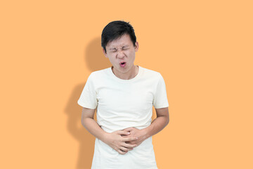 A young Asian man in a white t-shirt clutches his stomach against a orange background, expressing...