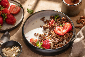 Healthy breakfast: fresh granola, muesli with milk, strawberry and almond for breakfast on brown textured background in bright morning light