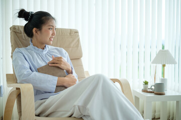Young woman sitting comfortably in a chair, hugging a book and smiling looks out the window.