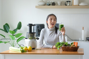 Cheerful woman enjoying a glass of fresh cold-pressed green vegetable juice in a bright kitchen....