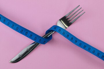Stainless fork tied with twisted blue measuring tape isolate on pink background. Diet and fit body...