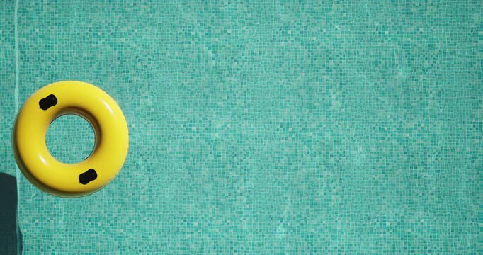 Top view of a bright yellow inflatable ring floating on clear turquoise water in a swimming pool. Sunlight casts a distinct shadow on the mosaic tiles. Minimal, peaceful summer vacation vibe