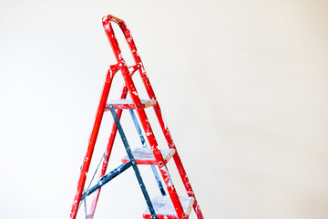 A red step ladder, set against a clean white wall, is ready for use. A red step ladder stands in...