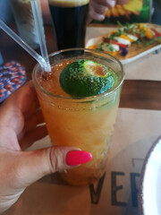 A close-up of a hand holding a cocktail glass with a lime slice. The drink is orange and served with two straws. A blurred background shows food and drinks.