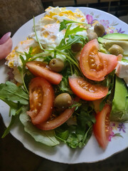 A fresh salad with arugula, sliced tomatoes, green olives, and a side of scrambled eggs on a floral plate. Concept of healthy eating and nutrition.