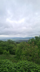 Obraz premium A scenic view of rolling hills under a cloudy sky. Lush green trees and shrubs dominate the foreground, creating a tranquil natural landscape.