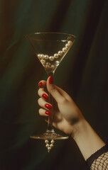 A hand with red nails holds up an  martini glass filled  with pearl necklace