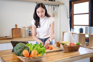Young woman preparing fresh vegetables in kitchen while following an online recipe on digital...