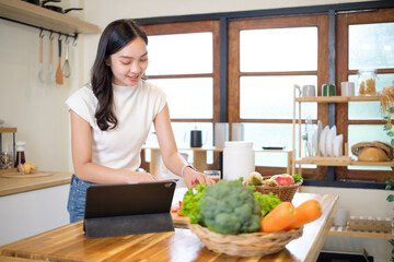 Young Asian woman preparing fresh vegetables while using a tablet in a cozy kitchen. Healthy eating and cooking at home concept.
