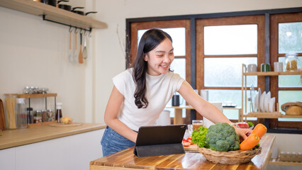 Smiling young woman in casual clothes preparing fresh vegetables in a cozy kitchen while following...