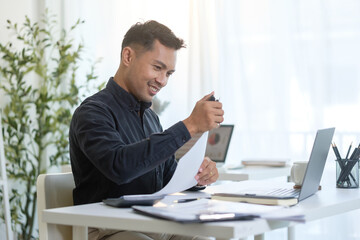 Smiling office worker organizing paperwork at his desk with a laptop and files.