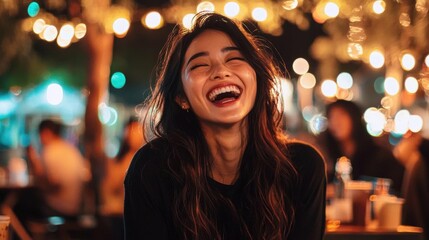 Close-up portrait of a young woman with long dark hair, laughing with her eyes closed and her mouth wide open. she is wearing a black top and appears to be in a restaurant or bar.